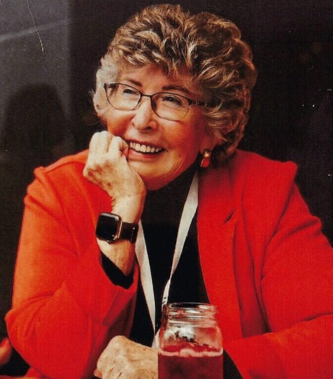 Smiling woman with curly hair wearing glasses and a red jacket.
